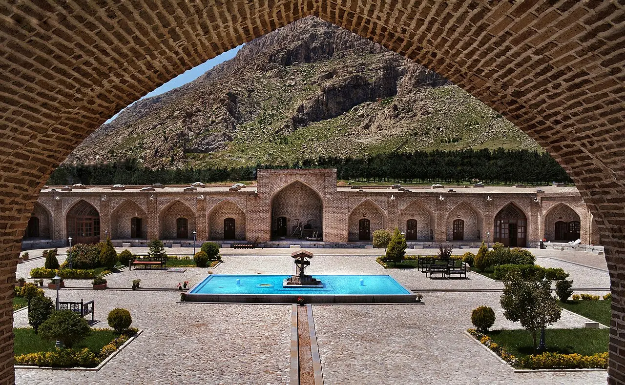 حیاط کاروانسرای شاه‌عباسی بیستون با حوض آبی که از میان یک طاق آجری دیده می‌شود و کوه در پس‌زمینه است