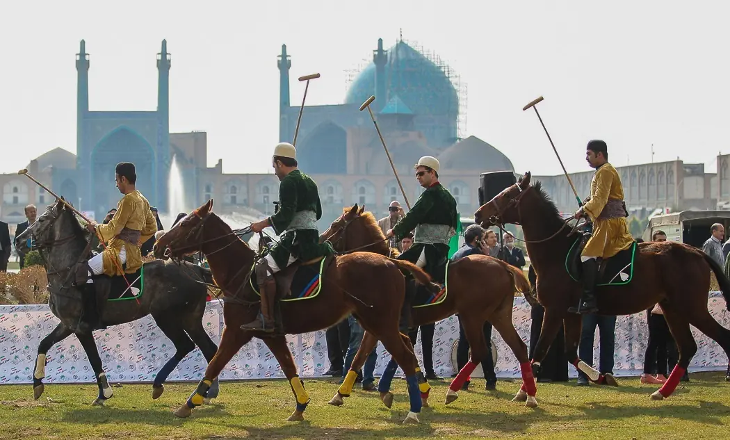 چوگان‌بازان با لباس سنتی سوار بر اسب در میدان نقش جهان با گنبد و مناره‌های مسجد امام در پس‌زمینه