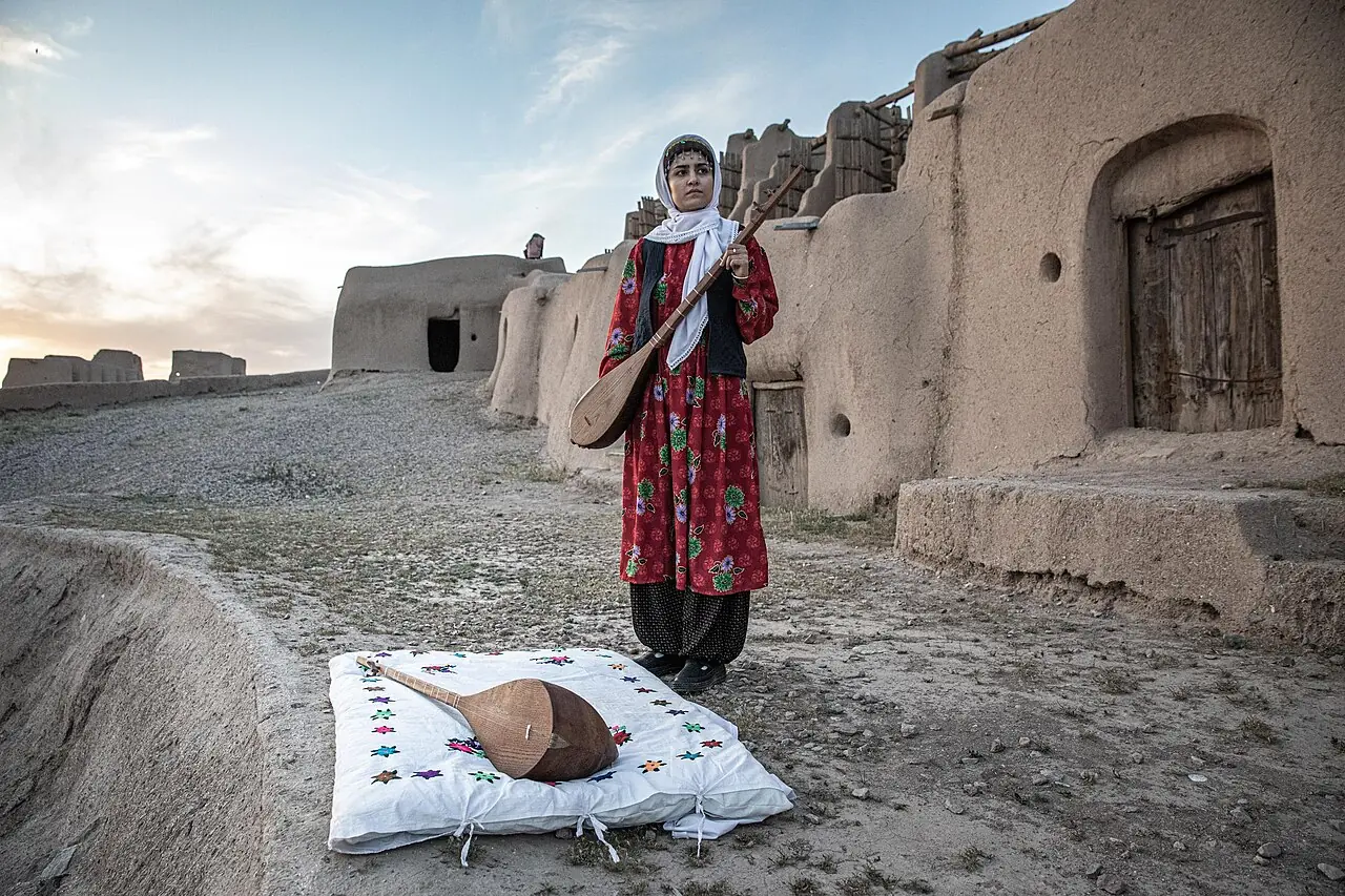 زنی با لباس محلی سرخ و روسری سفید در کوچه‌ای کاهگلی ایستاده و دوتار در دست دارد؛ دوتار دیگری روی پارچه‌ای جلوی اوست
