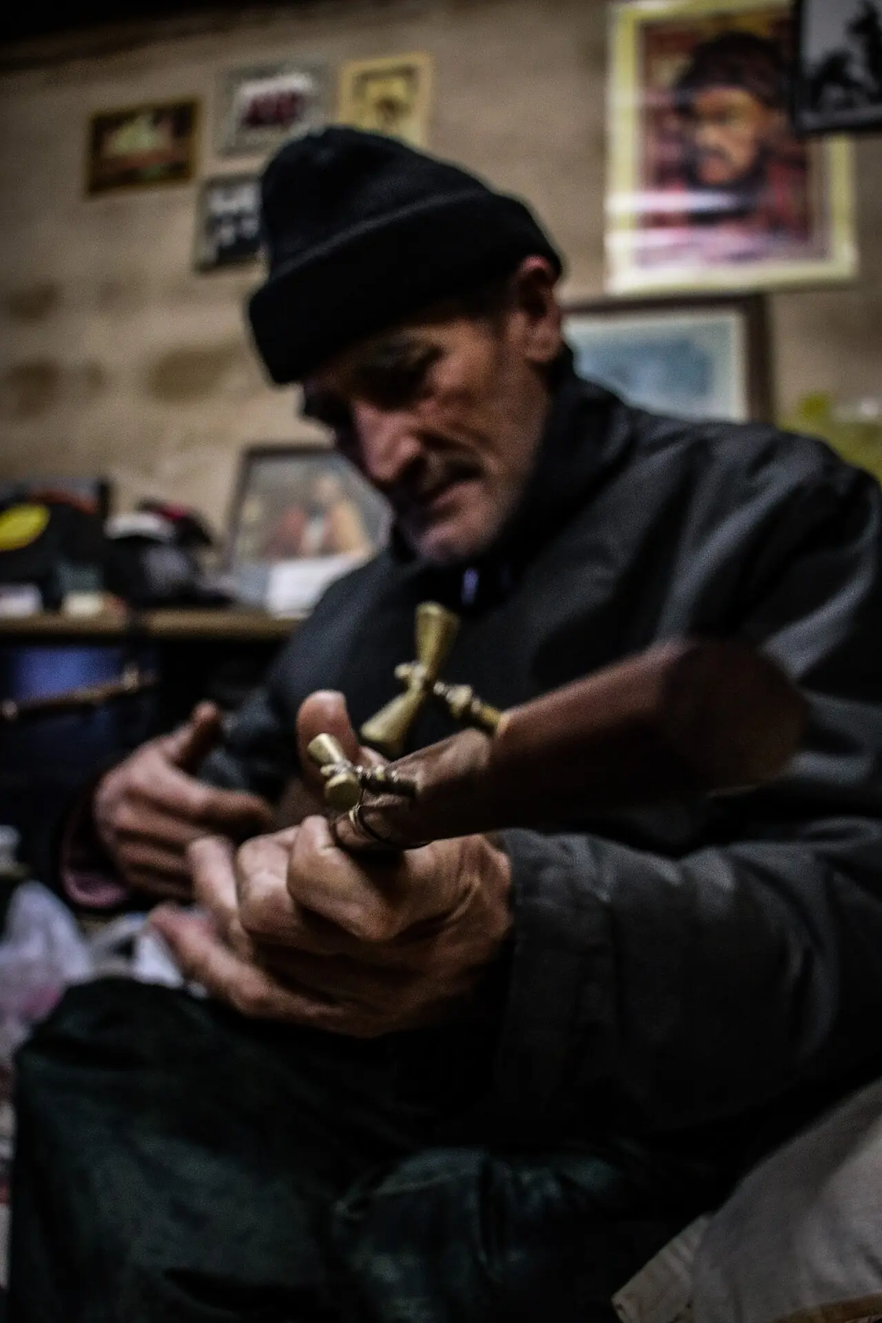 مردی ترکمن با کلاه بافتنی در کارگاهی پر از عکس، سر دوتار را در دست گرفته و کوک می‌کند