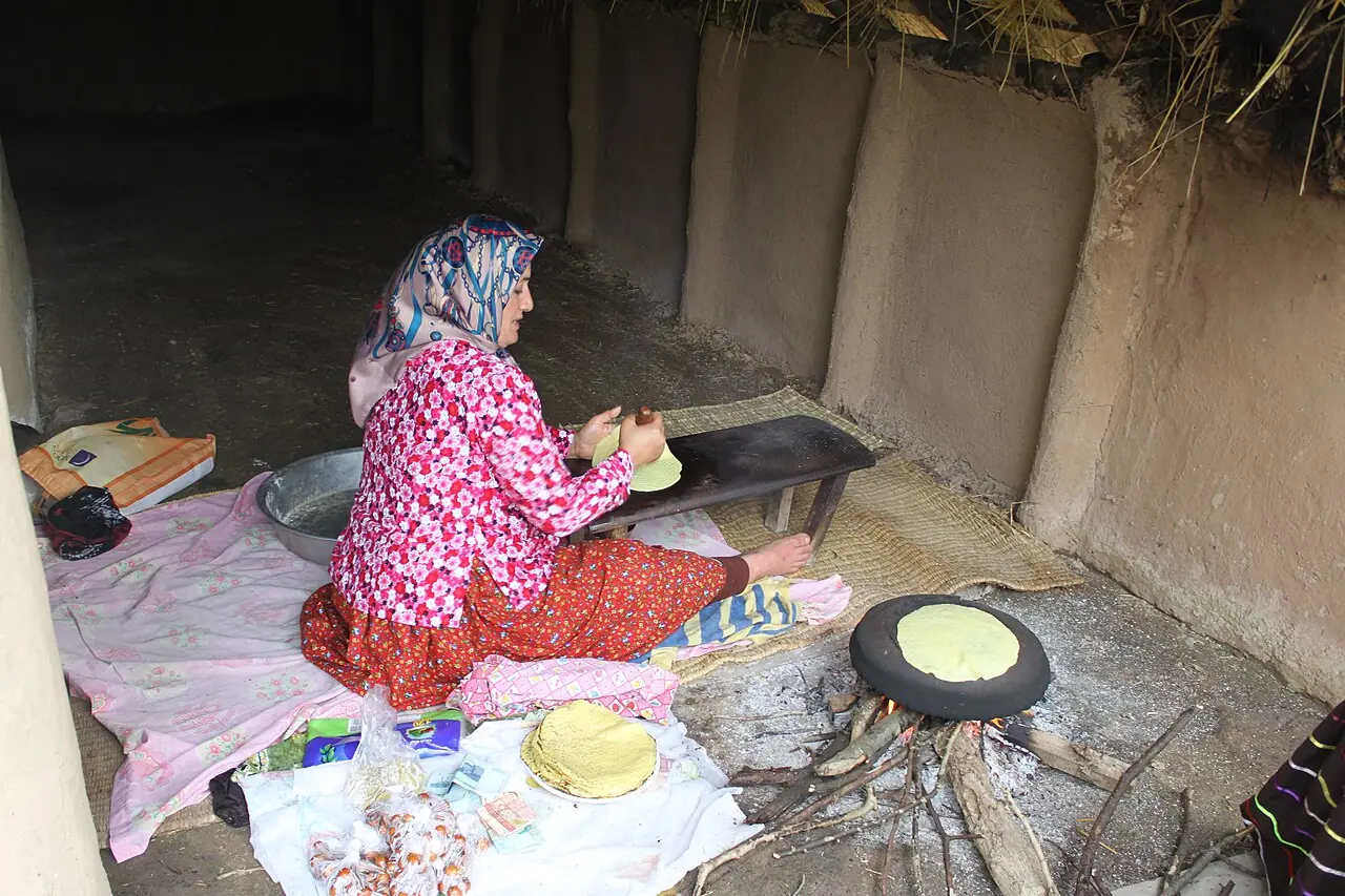 زنی با لباس محلی در کنار آتش هیزم نان نازک زردرنگی را روی تابه‌ای سیاه می‌پزد