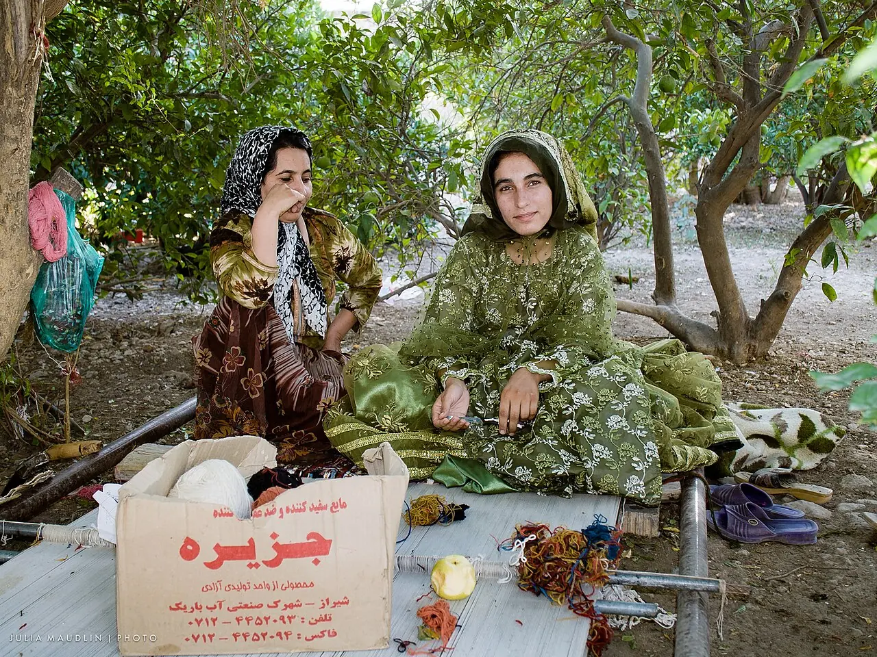 دو زن قشقایی با لباس محلی رنگارنگ کنار دار افقی قالی در سایه‌ی درختان نشسته‌اند