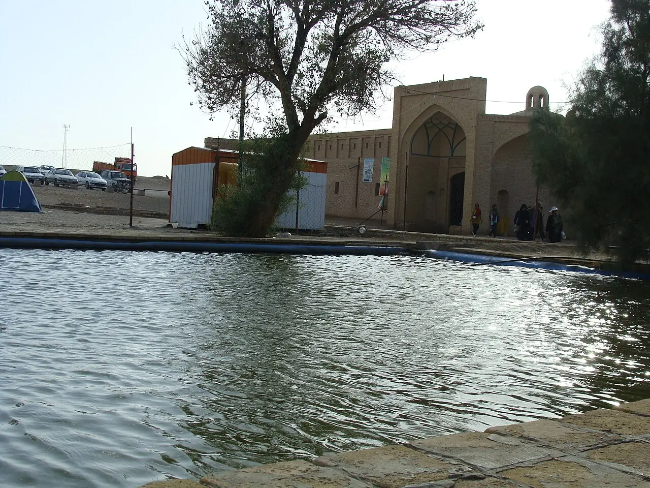 برکه‌ی آب کنار کاروانسرای مرنجاب با درختی کنار آن و سردر آجری کاروانسرا در پس‌زمینه