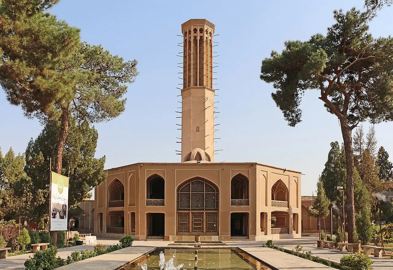 کوشک خشتی باغ دولت‌آباد با بادگیر بلند و حوض جلوی آن میان درختان کاج