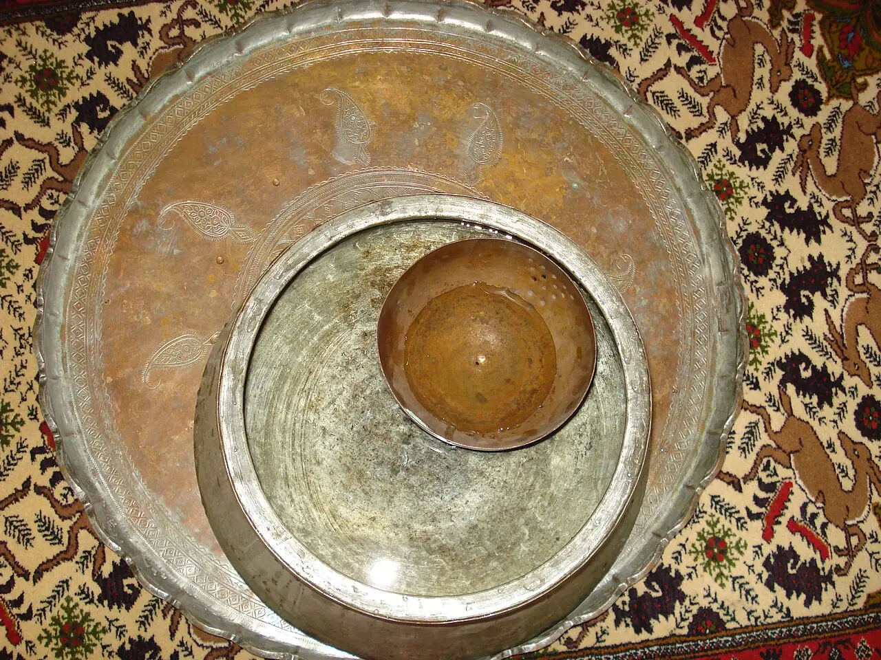 ساعت آبی سنتی با کاسه‌ای کوچک مسی درون ظرفی بزرگ‌تر روی فرش