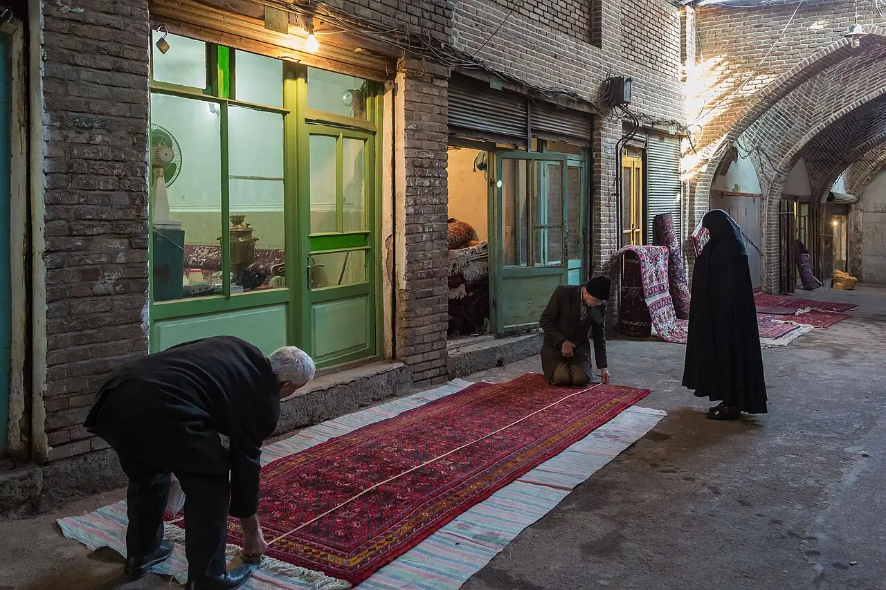 مردی در گذر بازار روی قالی پهن‌شده خم شده و آن را وارسی می‌کند