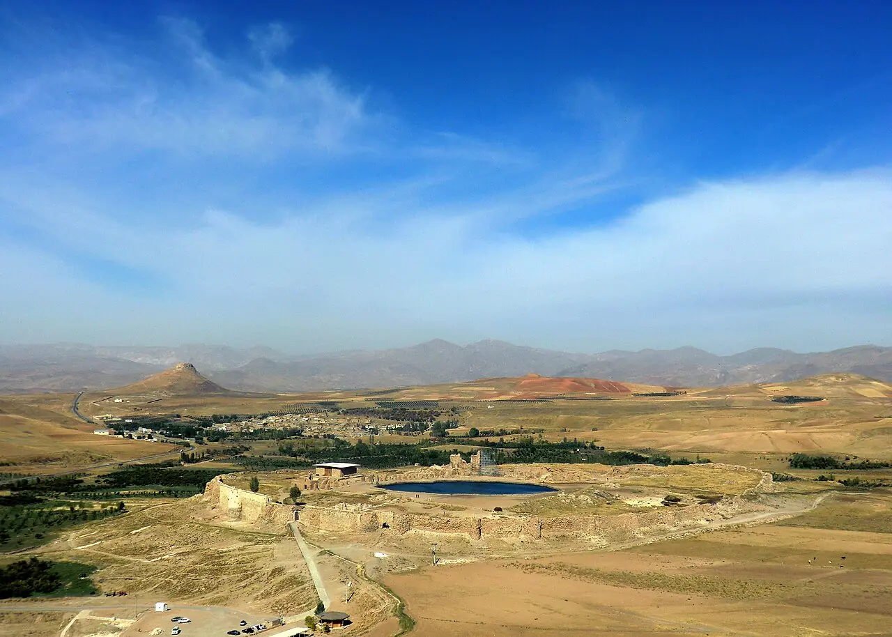 نمای هوایی تخت سلیمان با حصار بیضی‌شکل، دریاچه‌ی آبی وسط آن و دشت و کوه‌های اطراف